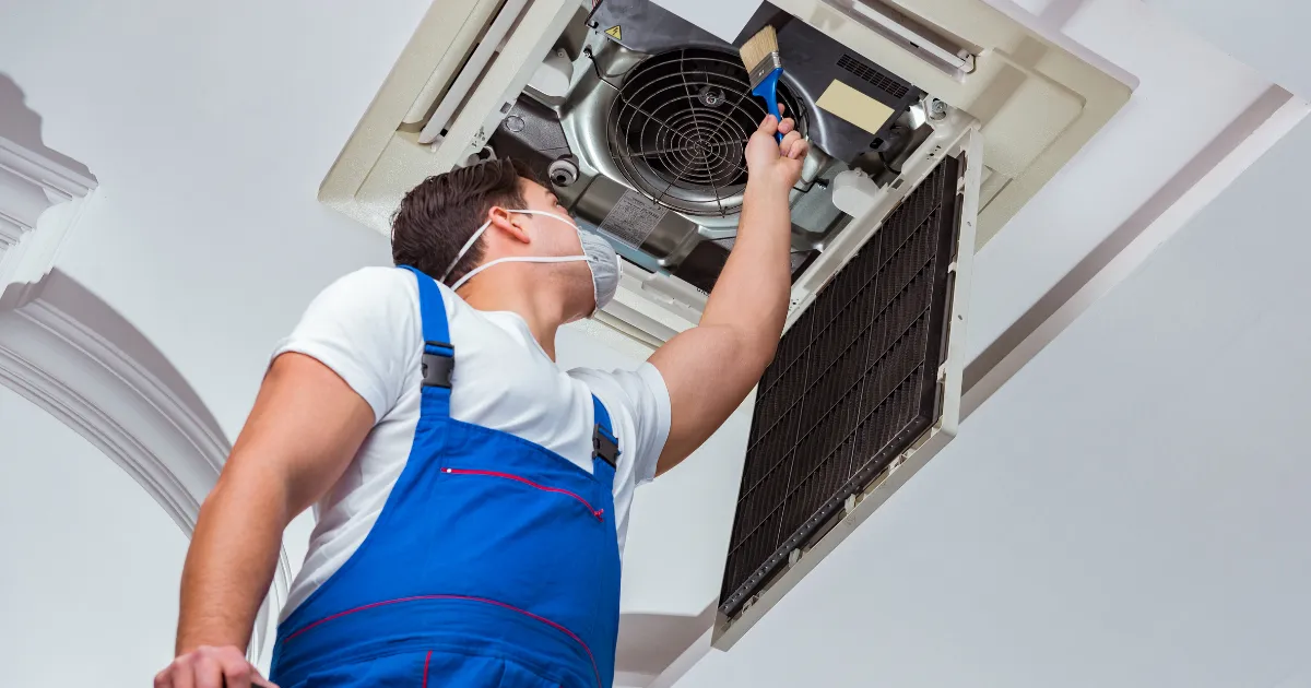 A Rapid Response team member is cleaning a ceiling-mounted AC unit with a brush to improve airflow and system efficiency