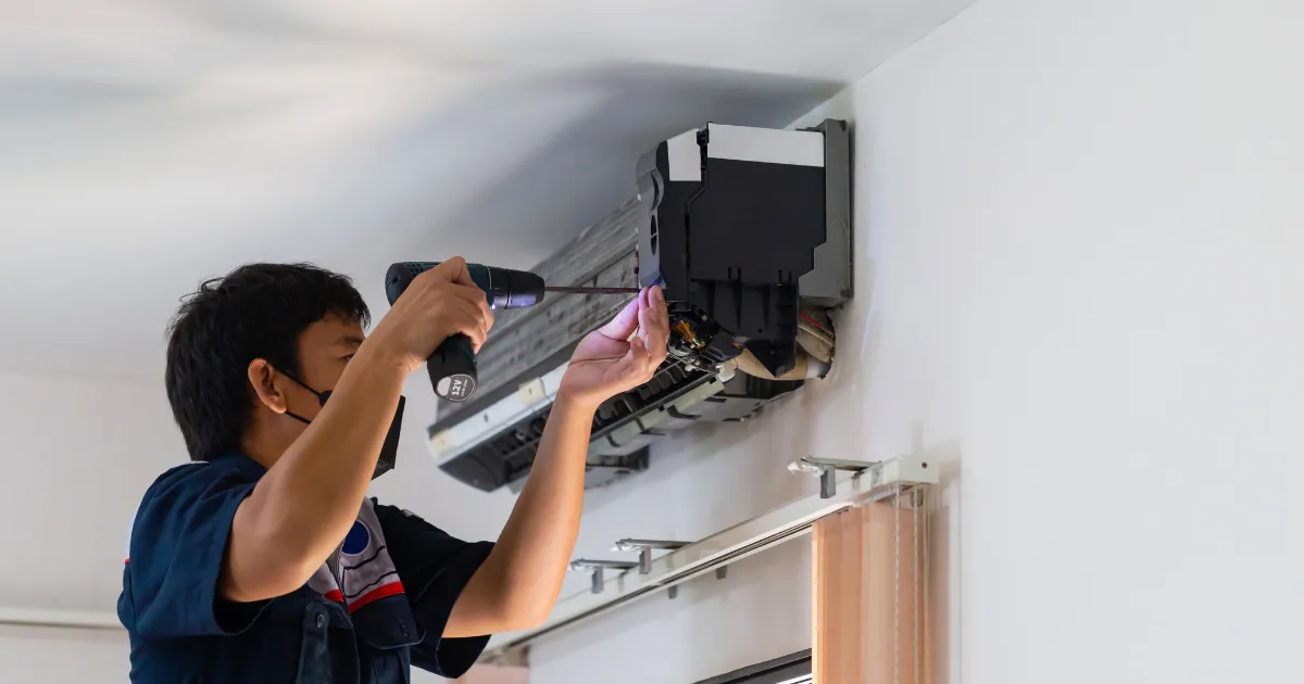 A technician uses a power drill to remove the front panel of a wall-mounted air conditioning unit during a repair or maintenance service