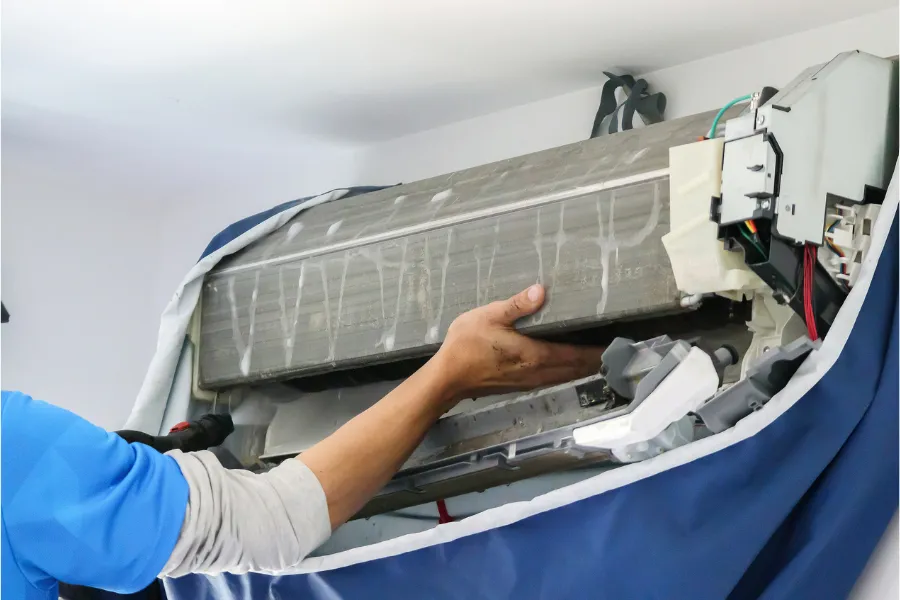 A technician carefully removes and cleans the evaporator coil of a wall-mounted AC unit during deep maintenance