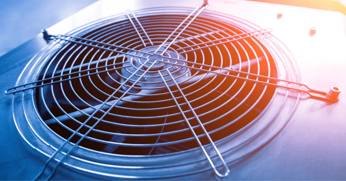 Close-up view of an outdoor HVAC condenser fan with a metal protective grille under warm and cool lighting