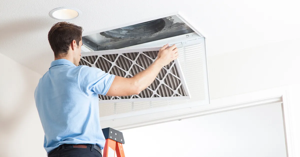 A person standing on a ladder while removing an old air filter from a ceiling vent