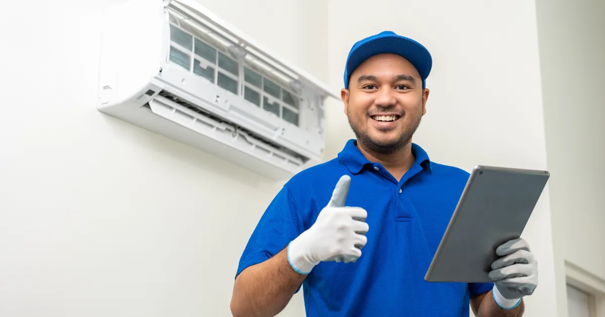 A technician standing in front of a wall-mounted air conditioner, holding a tablet and giving a thumbs up